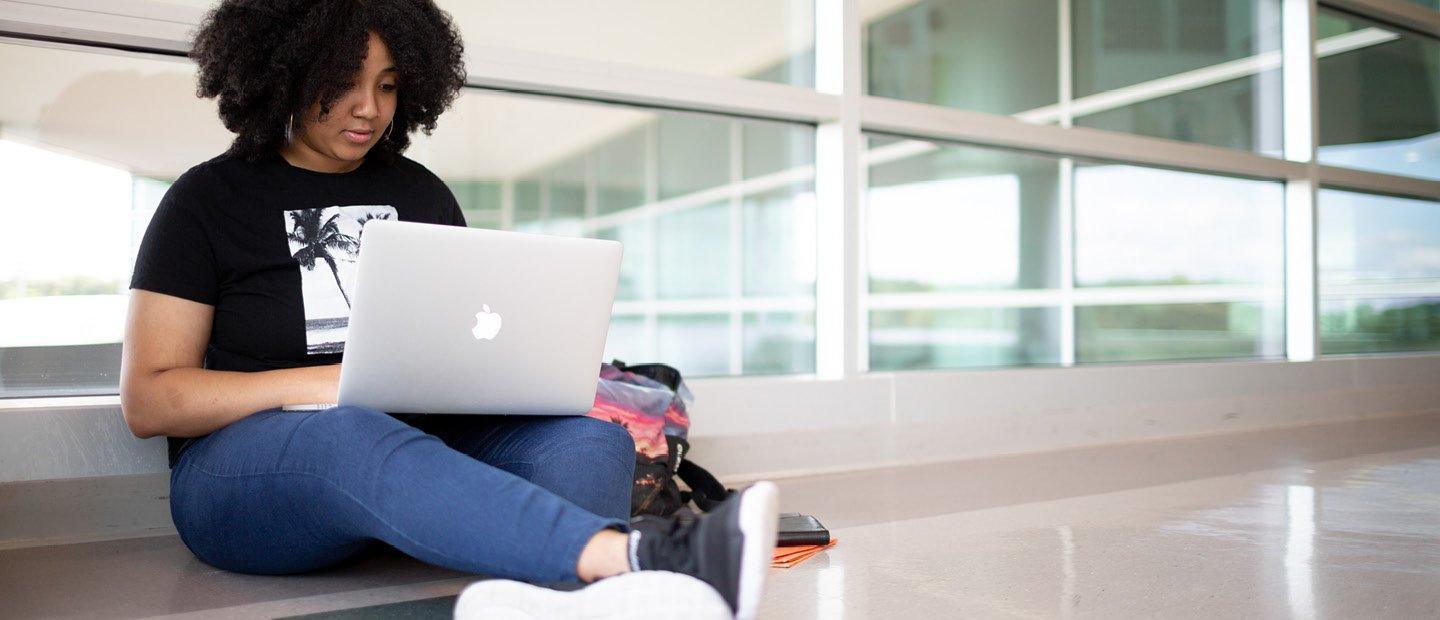 young woman seated on the floor in front of a window with an open laptop in her lap